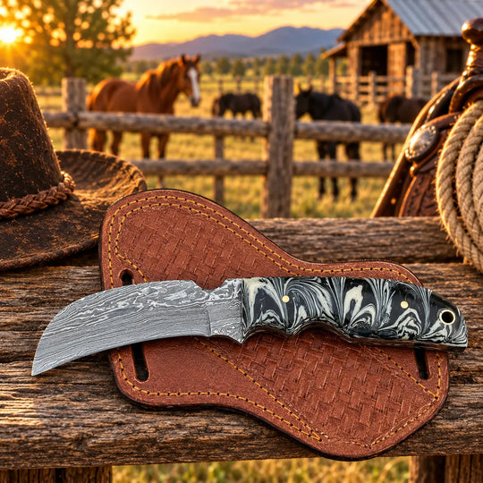 A folding pocket knife with a patterned black and white handle and Damascus steel blade rests on a brown woven leather sheath on a rustic wooden fence. The background shows a blurred outdoor ranch scene with horses, a barn, and mountains at sunset.