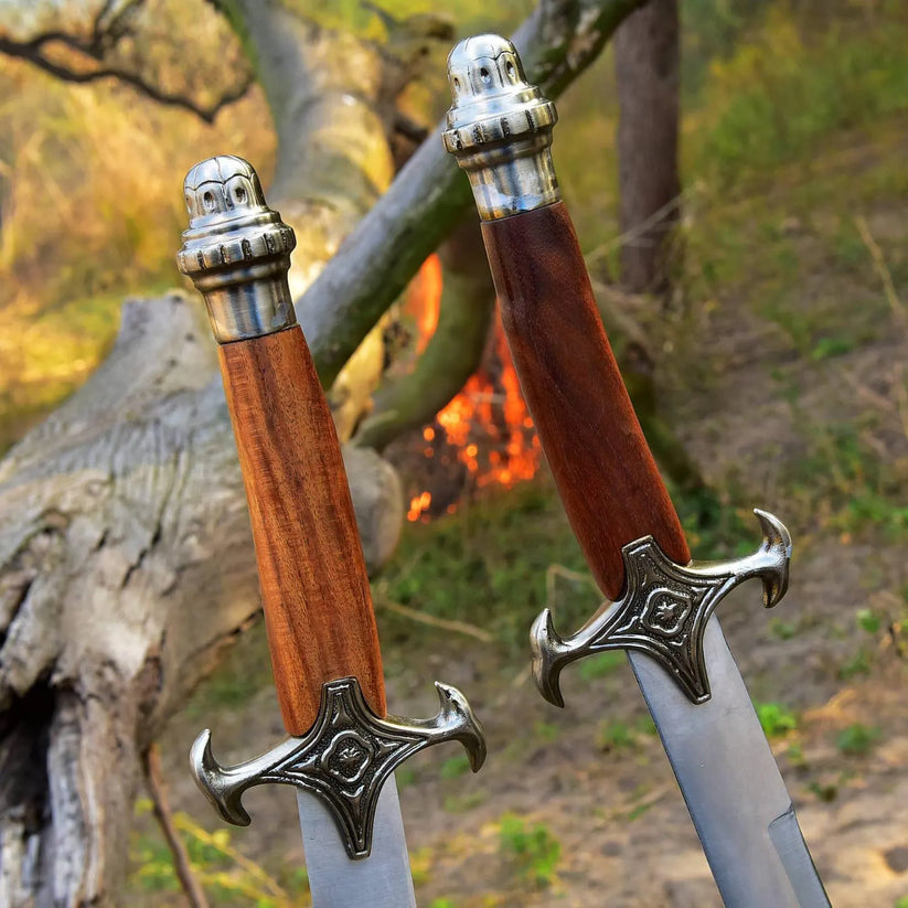 Two medieval-style swords with wooden handles and ornate silver blades are shown outdoors in a forest setting near a fire.