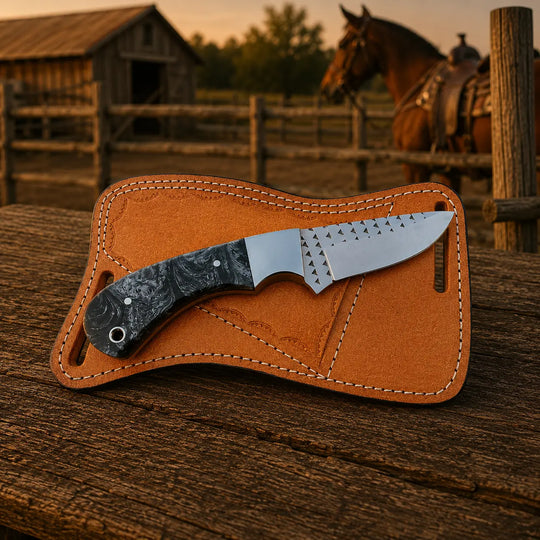 A fixed-blade knife with a dark marbled handle and a silver blade rests diagonally inside a tan leather sheath on a rustic wooden surface, with a barn, fence, and horse blurred in the background at sunset.
