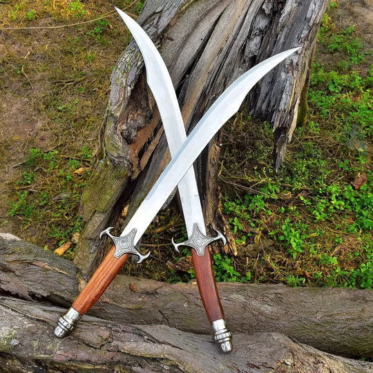 Two long curved swords with silver blades, silver crossguards, and brown wooden handles are crossed against a weathered tree trunk outdoors with grass and soil visible.