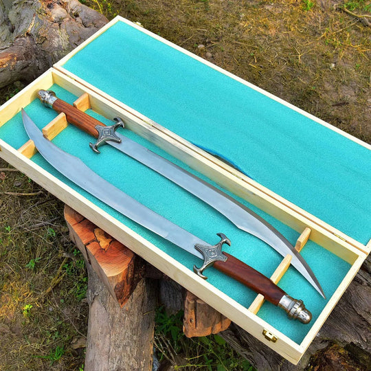 A decorative sword with a curved silver blade, brown wooden handle, and ornate silver guard is displayed inside an open light wood presentation box lined with turquoise fabric, set outdoors on a log with trees and grass in the background.