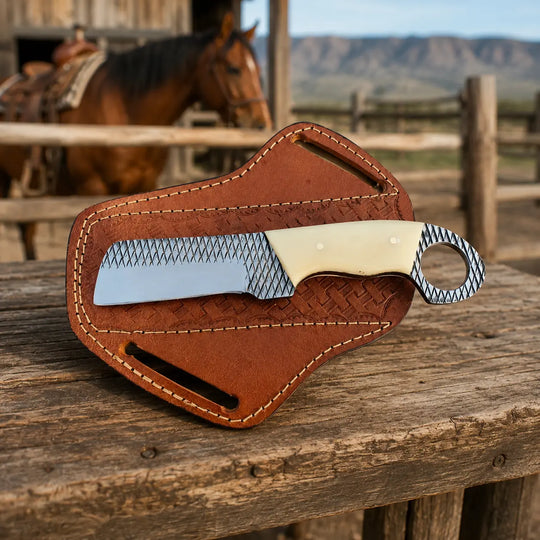A fixed-blade knife with a textured silver blade and cream handle rests on a brown leather sheath, placed on a rustic wooden table with a blurred western ranch background featuring a horse and mountains.