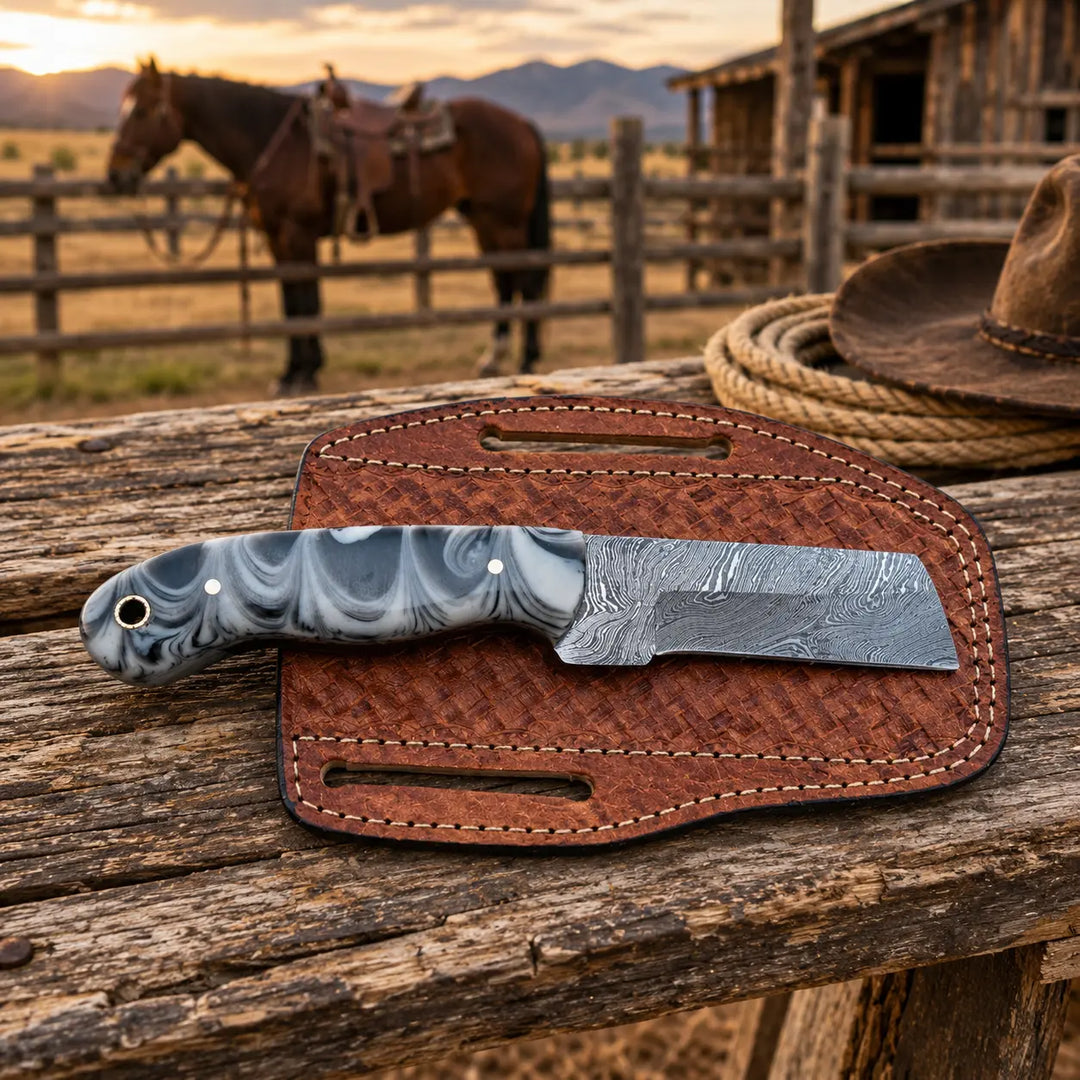 A custom knife with a marbled gray and white handle and a Damascus steel blade rests on its brown textured sheath, placed on a rustic wooden table with a blurred background of a horse, barn, and mountains at sunset.