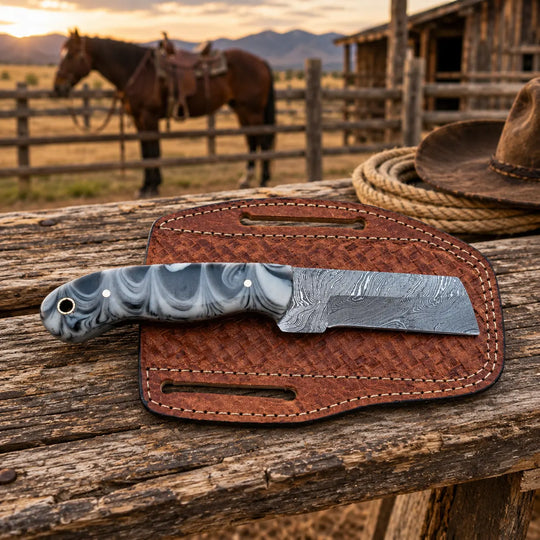 A custom knife with a marbled gray and white handle and a Damascus steel blade rests on its brown textured sheath, placed on a rustic wooden table with a blurred background of a horse, barn, and mountains at sunset.