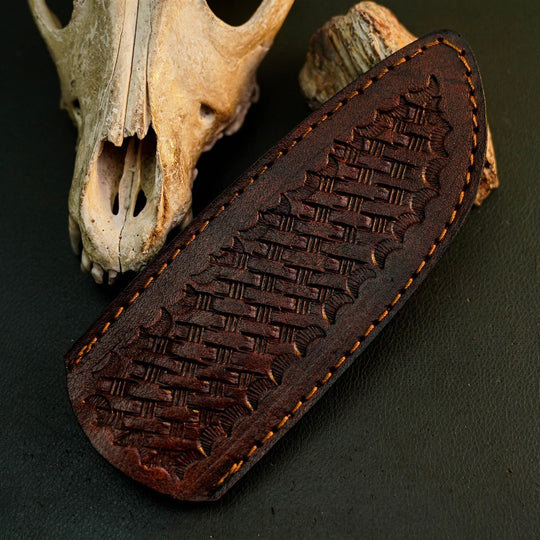 A dark brown leather knife sheath with an embossed basketweave pattern and tan stitching, displayed on a dark textured surface next to a dried animal skull.
