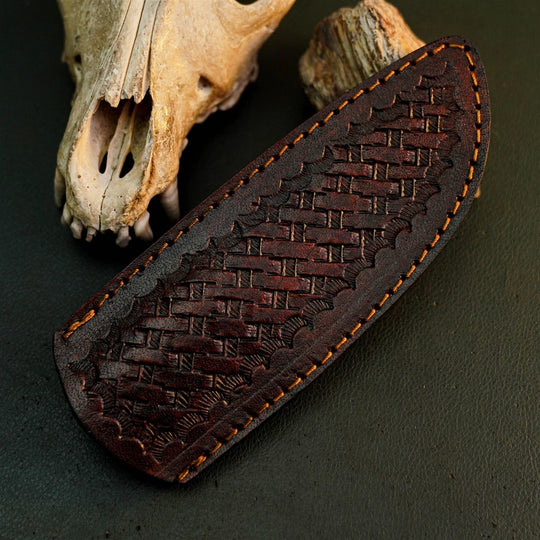 A dark brown leather sheath with an embossed basketweave pattern and orange-brown contrast stitching, displayed on a dark textured surface next to animal bones.