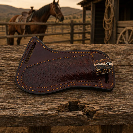 A dark brown leather knife sheath with a textured basketweave pattern and orange contrast stitching, resting on a rustic wooden table with a blurred western background showing a horse and a barn.