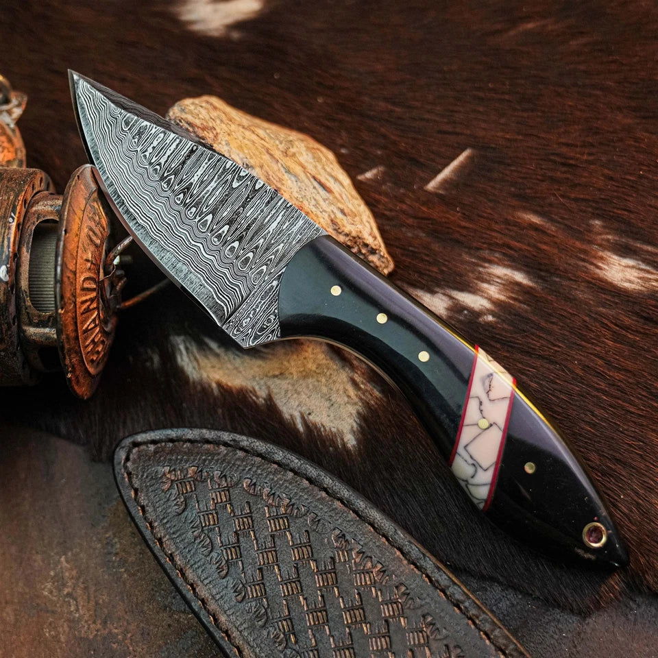 A close-up of a fixed-blade knife with a patterned steel blade, black handle, and decorative inlay, resting on a brown and white cowhide surface with a leather sheath and rustic props nearby.
