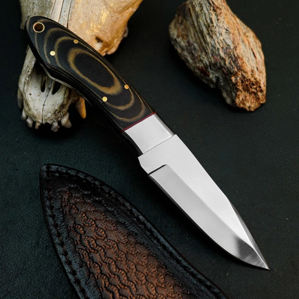 A fixed-blade knife with a polished silver blade and a black handle featuring gold swirls and brass pins, displayed diagonally on a dark textured surface with a leather sheath, a bone skull, and a rock.