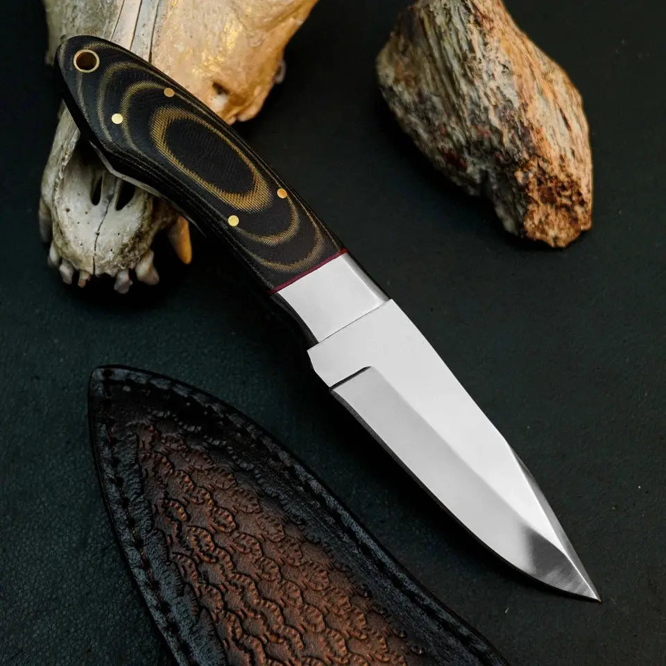 A fixed-blade knife with a polished silver blade and a black handle featuring gold swirls and brass pins, displayed diagonally on a dark textured surface with a leather sheath, a bone skull, and a rock.