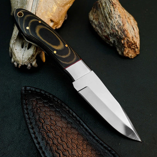 A fixed-blade knife with a polished silver blade and a black handle featuring gold swirls and brass pins, displayed diagonally on a dark textured surface with a leather sheath, a bone skull, and a rock.