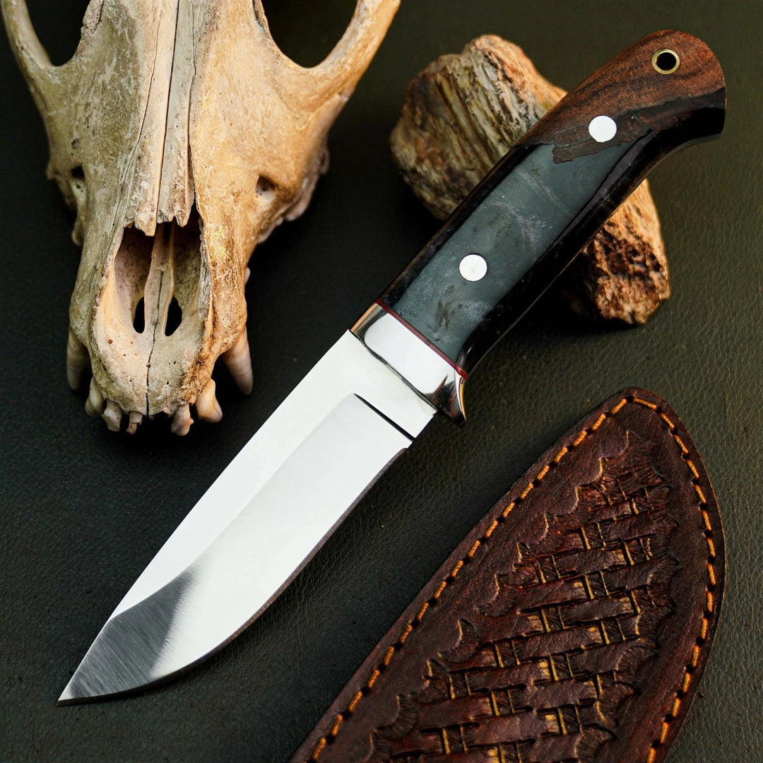 A fixed-blade knife with a polished silver blade, dark wood handle, and black inlay, displayed next to its brown leather sheath on a dark textured surface with a skull and wood props.