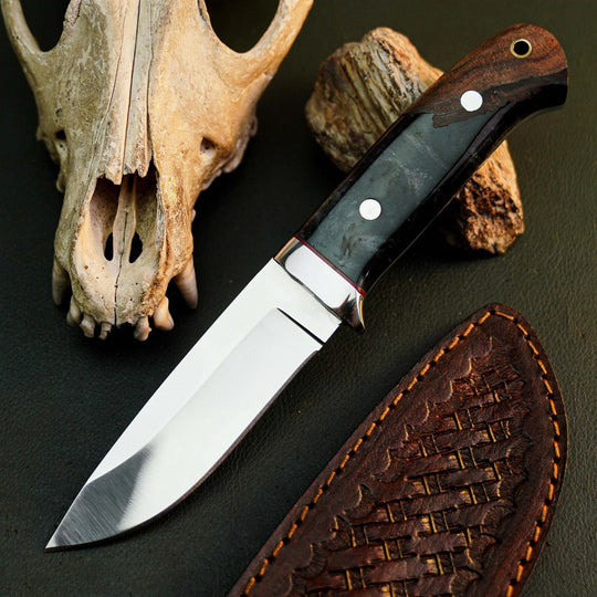 A fixed-blade knife with a polished silver blade, dark wood handle, and black inlay, displayed next to its brown leather sheath on a dark textured surface with a skull and wood props.
