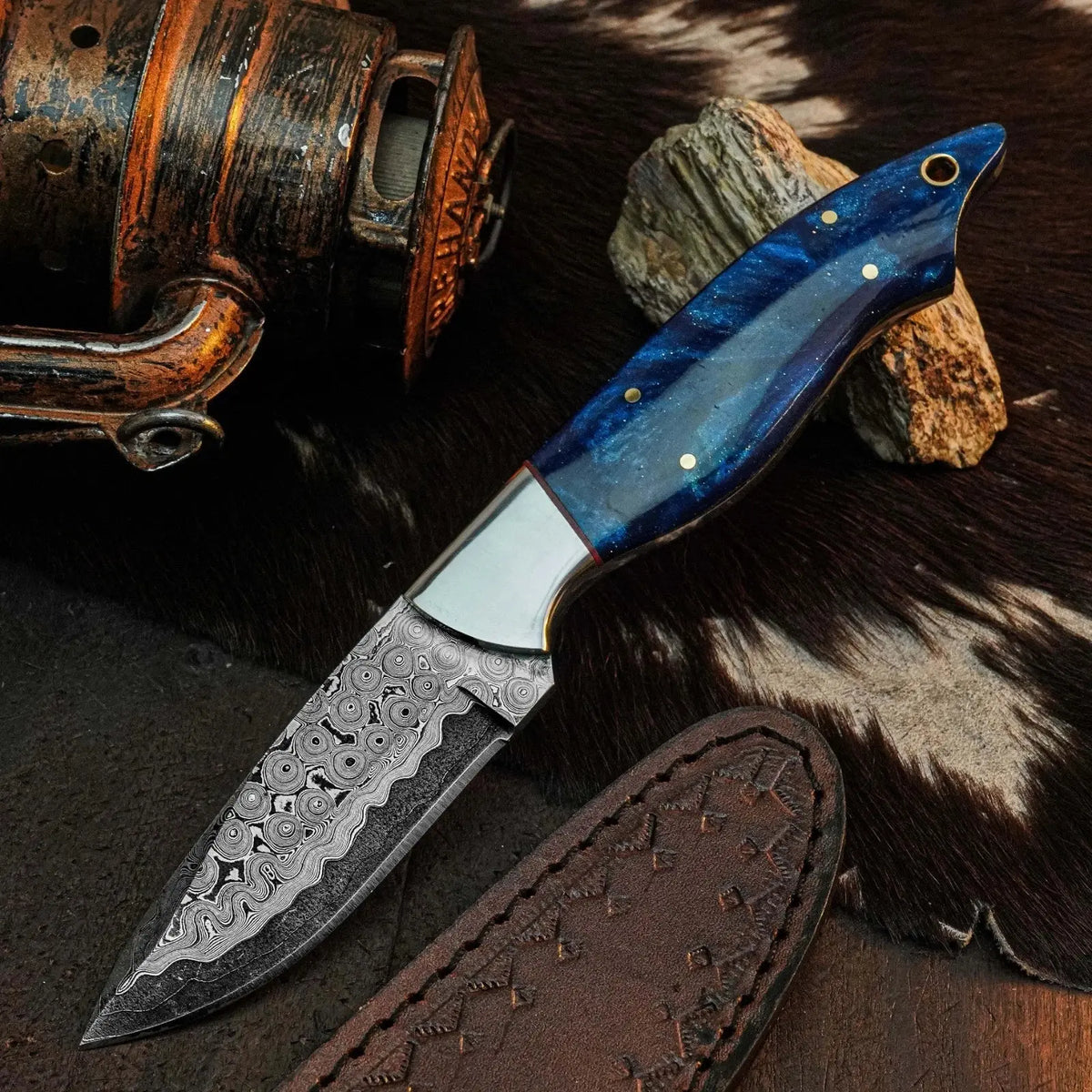 A fixed-blade knife with a patterned Damascus steel blade and a blue marbled wood handle, displayed on a dark textured surface with a leather sheath, rustic metal object, and cowhide background.