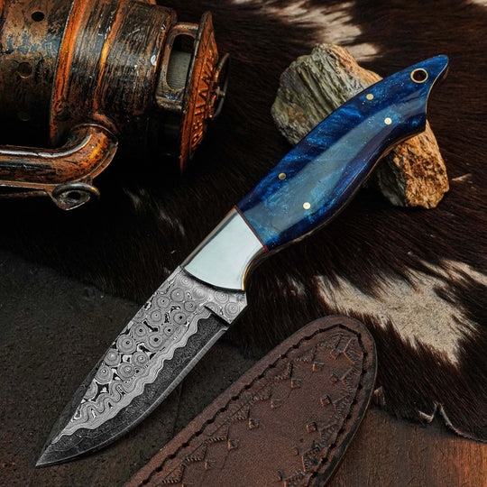 A fixed-blade knife with a patterned Damascus steel blade and a blue marbled wood handle, displayed on a dark textured surface with a leather sheath, rustic metal object, and cowhide background.