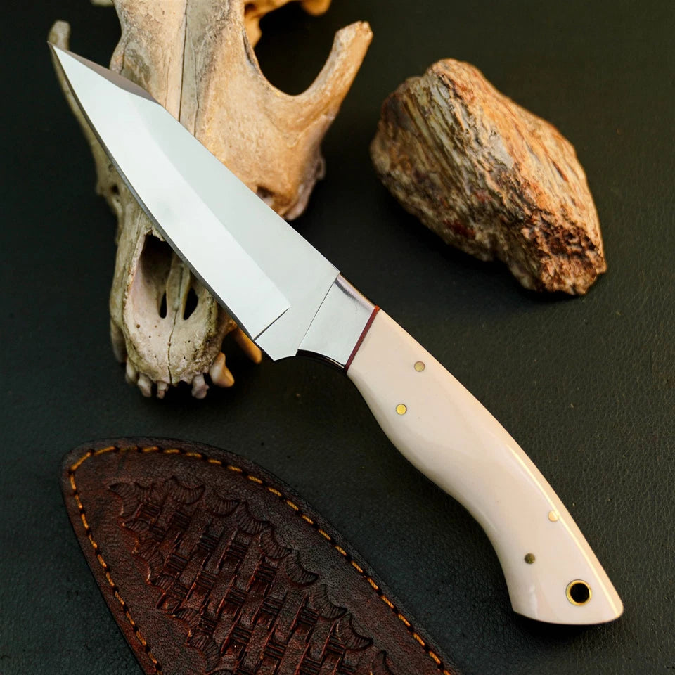A fixed-blade knife with a satin-finished silver blade and a cream-colored handle with brass pins, displayed diagonally on a dark textured surface next to a brown leather sheath, a deer skull, and a rock.