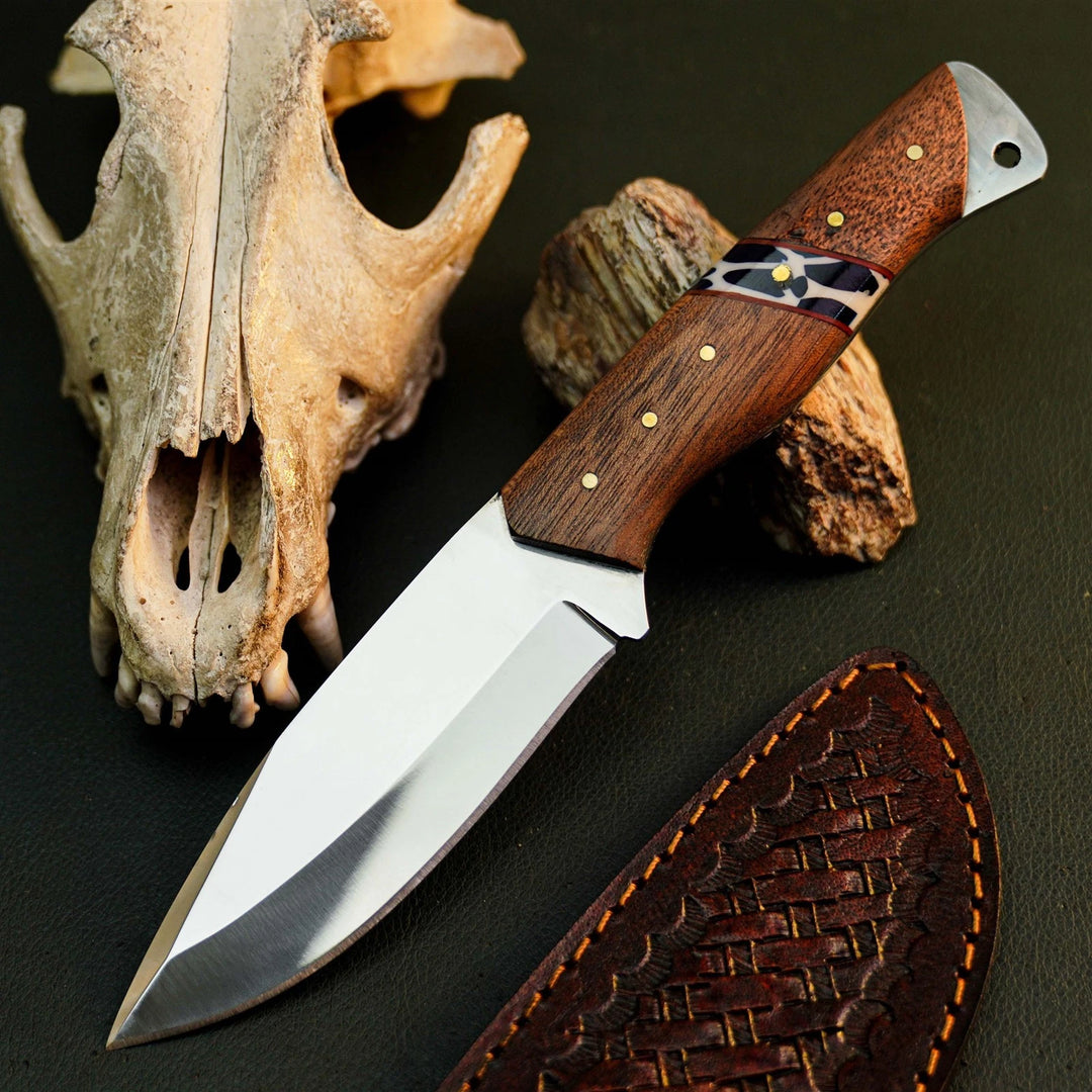 A fixed-blade knife with a polished silver blade and a brown wood-grain handle with brass pins, displayed diagonally on a dark textured surface next to a carved bone skull, a piece of driftwood, and a brown woven sheath.