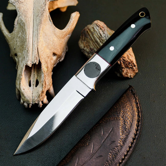 A fixed-blade knife with a satin-finished silver blade and a black handle with silver rivets, displayed diagonally on a dark textured surface alongside a leather sheath, a bone skull, and a piece of wood.