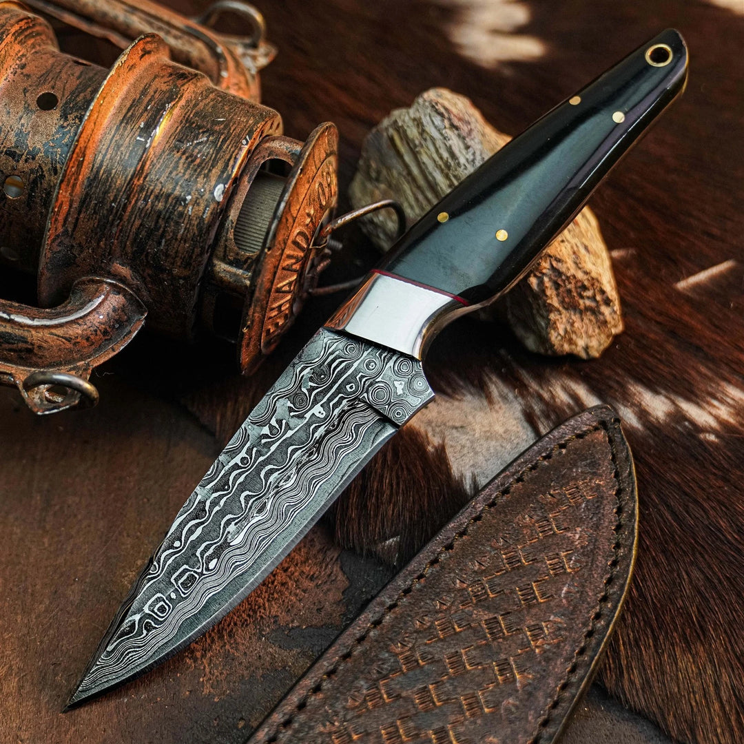 A fixed-blade knife with a patterned Damascus-style steel blade, a polished silver bolster, and a glossy black handle with brass pins, displayed diagonally on a textured brown surface next to a leather sheath and rustic wooden props.