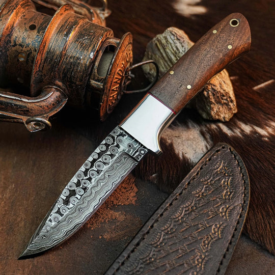 A fixed-blade knife with a patterned Damascus steel blade, a white bolster, and a dark brown wooden handle, displayed diagonally with its matching embossed brown leather sheath on a rustic brown textured background.