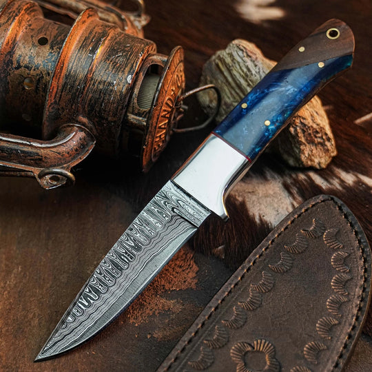 A fixed-blade knife with a patterned Damascus steel blade and a blue marbled resin handle, displayed diagonally next to its brown leather sheath on a rustic wooden surface with a fishing reel and stones.