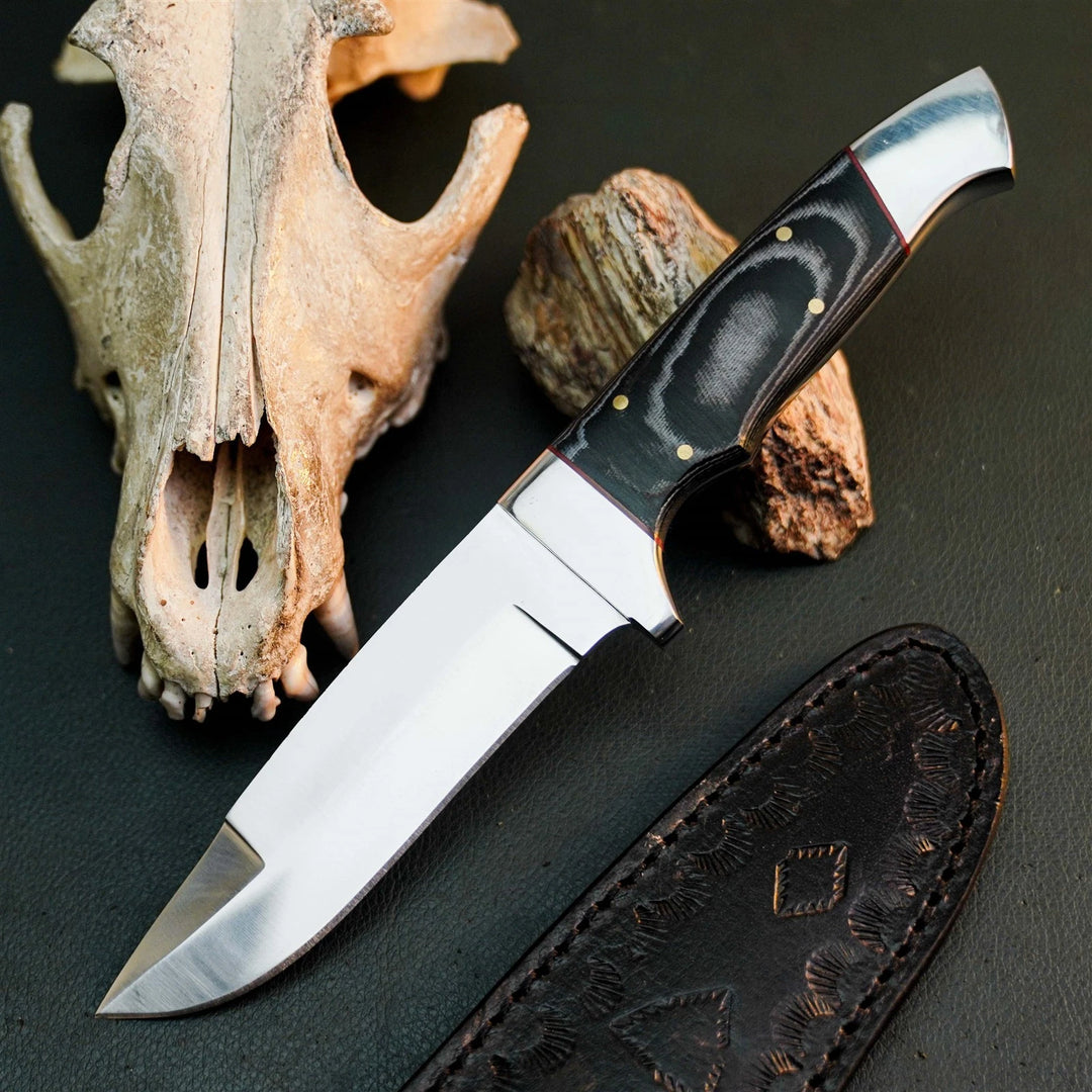 A fixed-blade knife with a polished silver blade and a black handle with gold pins, displayed diagonally on a dark textured surface next to a leather sheath, a bone skull, and a piece of wood.