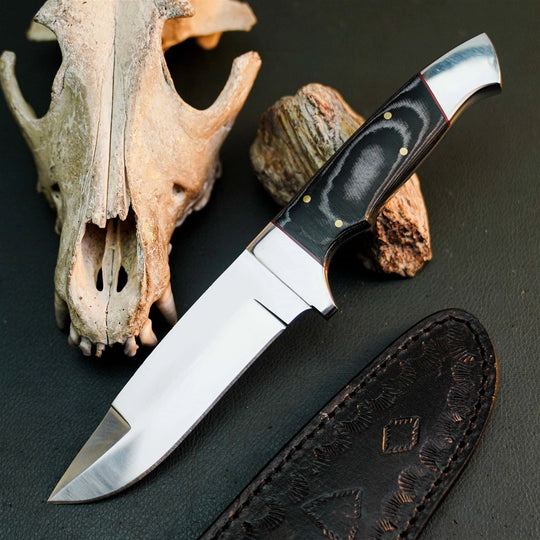 A fixed-blade knife with a polished silver blade and a black handle with gold pins, displayed diagonally on a dark textured surface next to a leather sheath, a bone skull, and a piece of wood.