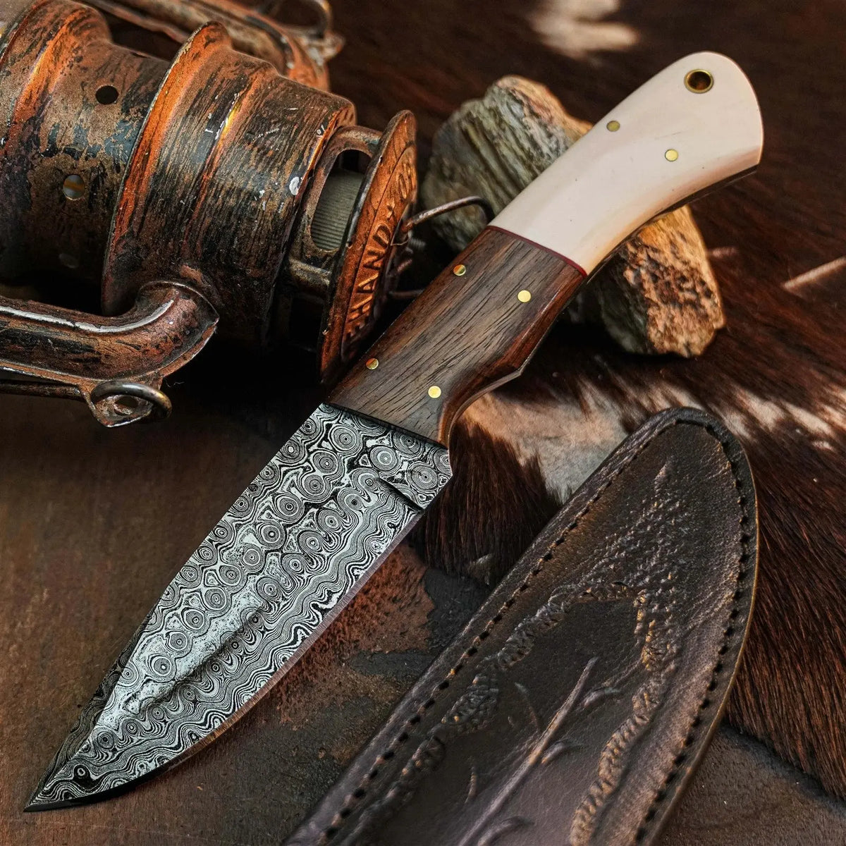 A fixed-blade knife with a patterned Damascus steel blade, a dark wood handle, and a cream-colored bone or horn handle section, displayed next to its dark brown leather sheath on a rustic background with a fishing reel and wood.