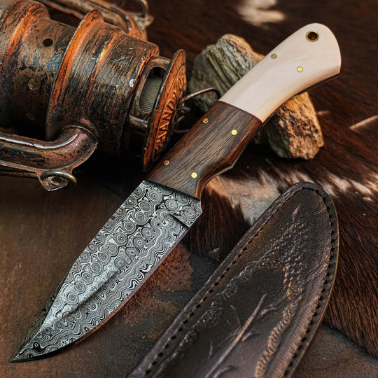 A fixed-blade knife with a patterned Damascus steel blade, a dark wood handle, and a cream-colored bone or horn handle section, displayed next to its dark brown leather sheath on a rustic background with a fishing reel and wood.