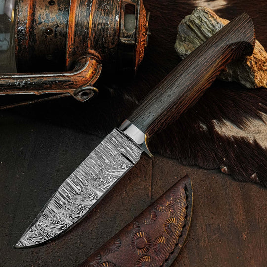A custom fixed-blade knife with a patterned Damascus steel blade and a dark wood handle, displayed diagonally on a rustic dark surface next to its leather sheath.