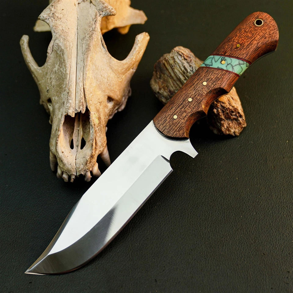 A fixed-blade knife with a polished silver blade and a brown wood handle featuring a turquoise inlay, displayed on a dark textured surface next to a bone skull.