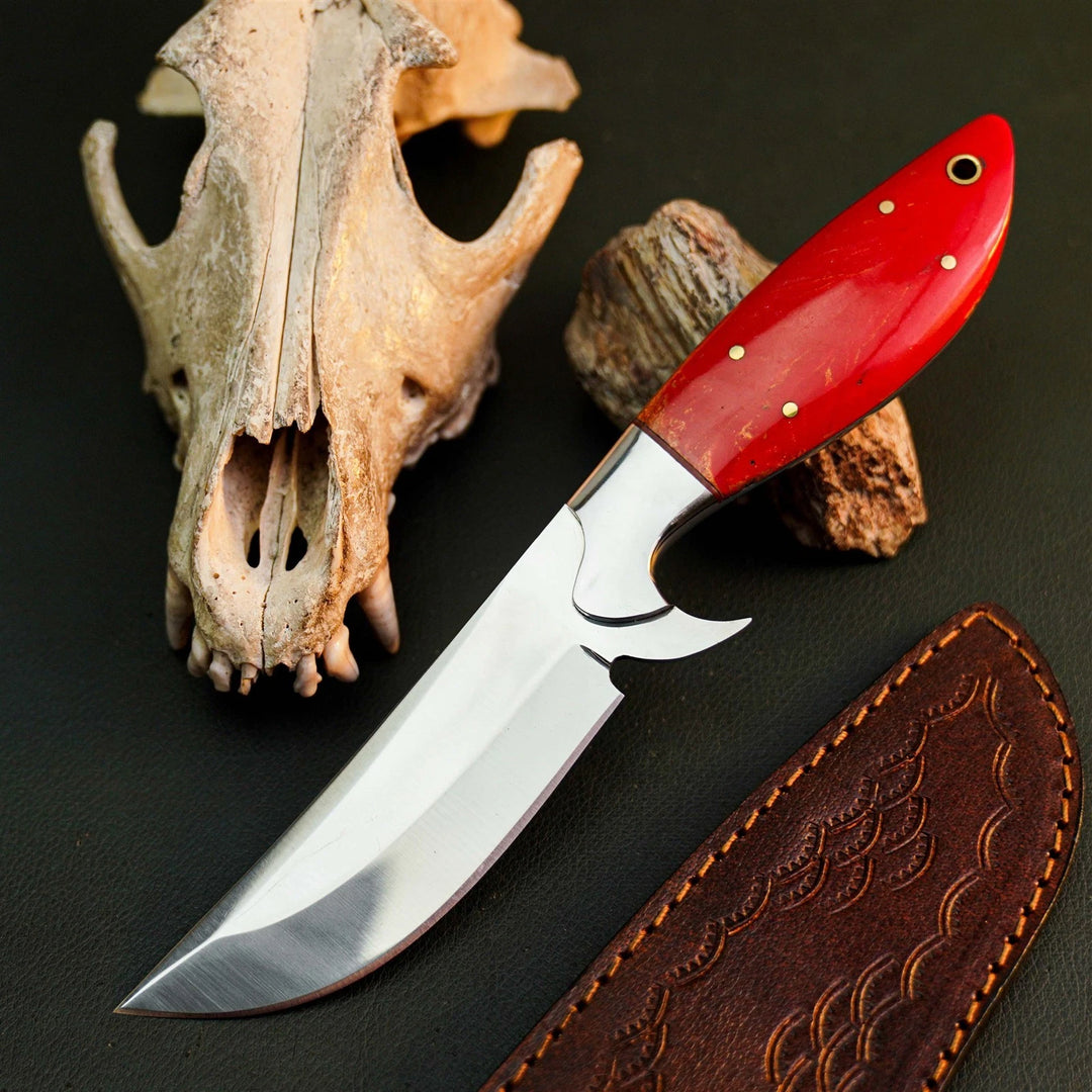 A fixed-blade knife with a polished silver blade and a bright red handle, arranged on a dark surface with a carved bone skull, small rocks, and a brown leather sheath.