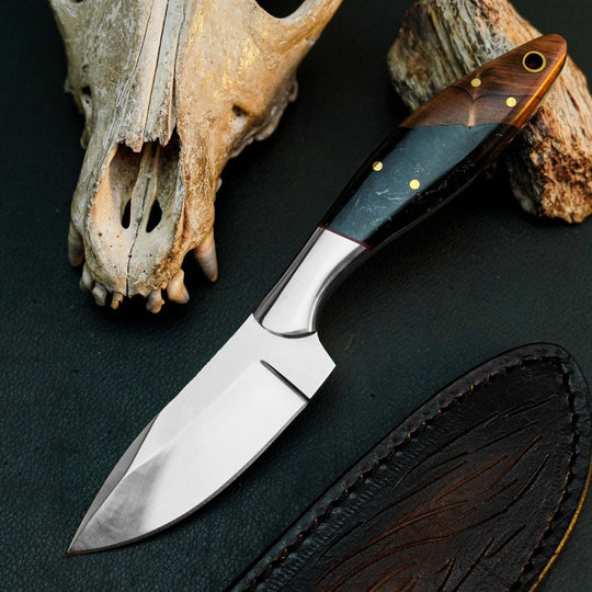 A fixed-blade knife with a satin-finished silver blade and a dark wood-and-stone handle, displayed diagonally on a dark textured surface next to a leather sheath, a deer skull, and a piece of wood.
