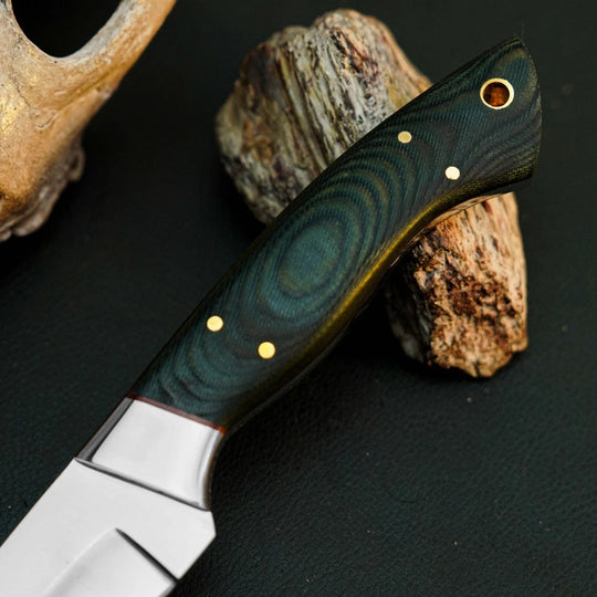 A close-up of a knife with a dark green and black patterned handle and brass pins, resting on natural wood pieces against a dark background.