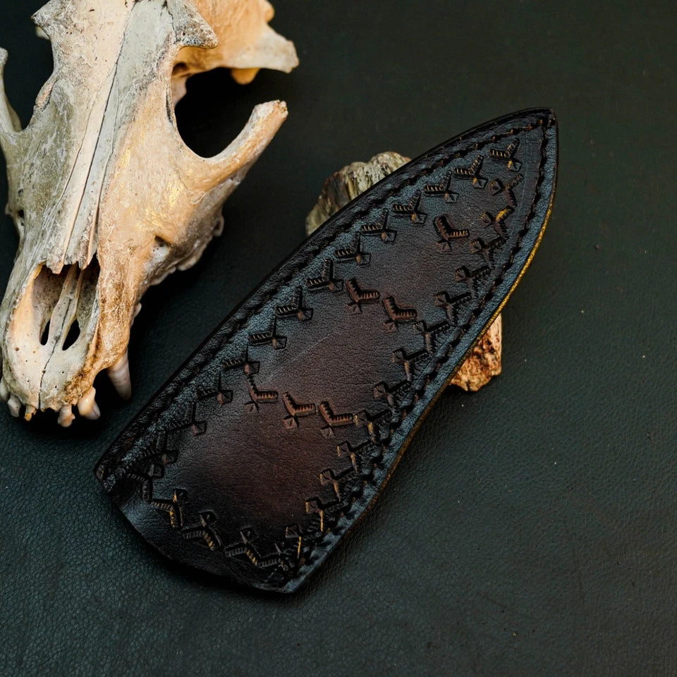A dark brown leather knife sheath with decorative stitched patterns, displayed on a dark textured surface next to a weathered animal skull.