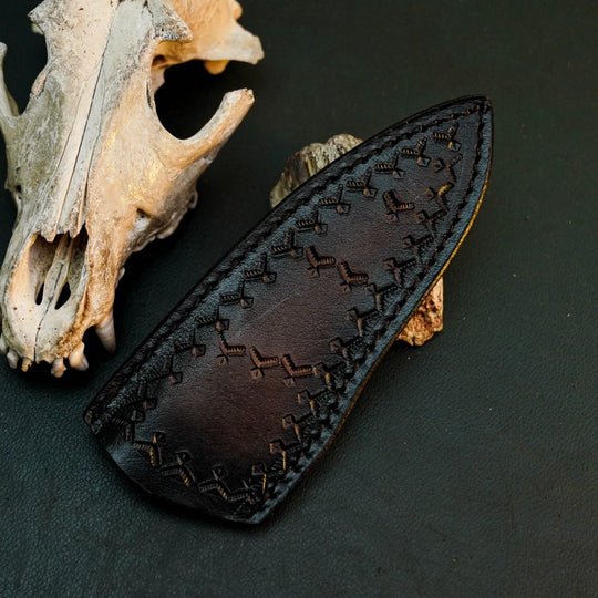 A dark brown leather knife sheath with decorative stitched patterns, displayed on a dark textured surface next to a weathered animal skull.