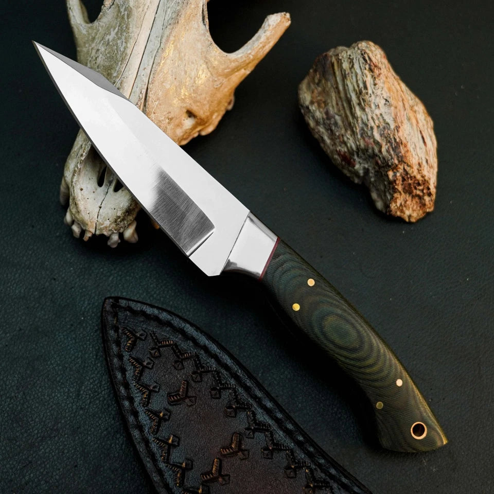 A fixed-blade knife with a satin-finished silver blade and a dark green wood-grain handle with brass pins, displayed diagonally on a dark textured surface with a stitched black sheath, a bone antler prop, and a rock.