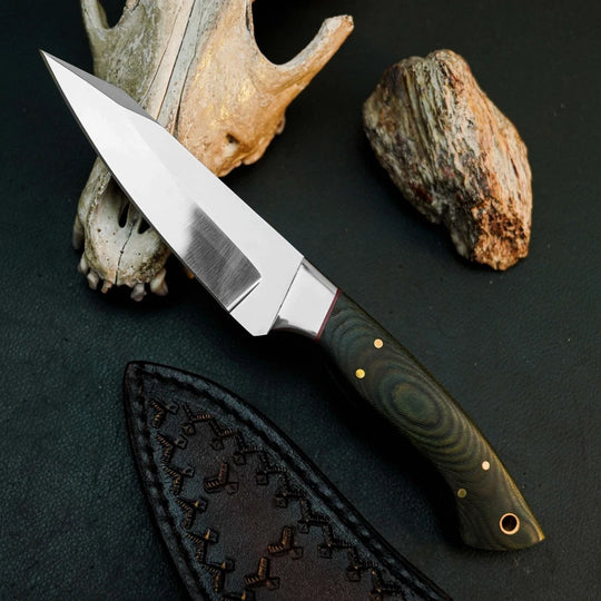 A fixed-blade knife with a satin-finished silver blade and a dark green wood-grain handle with brass pins, displayed diagonally on a dark textured surface with a stitched black sheath, a bone antler prop, and a rock.