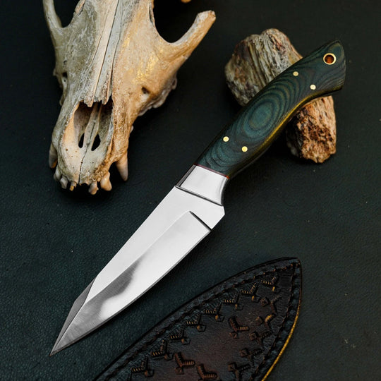 A fixed-blade knife with a polished silver blade and a dark green wood-grain handle with brass pins, displayed diagonally on a dark textured surface next to a leather sheath, a deer skull, and rocks.