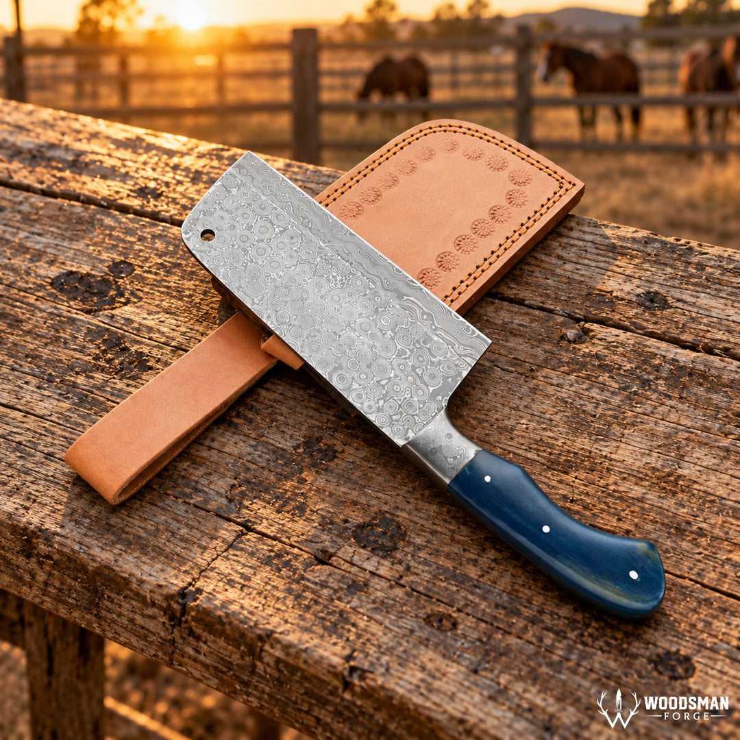 A cleaver-style knife with a patterned steel blade, blue handle, and tan leather sheath resting on a rustic wooden table outdoors at sunset, with horses visible in the background.