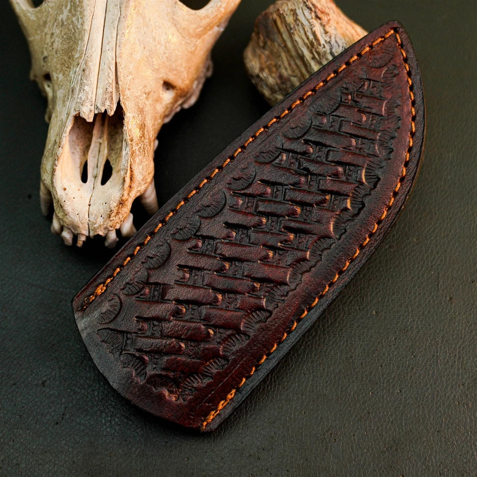 A dark brown leather knife sheath with embossed fish patterns and orange stitching, displayed on a dark textured surface next to animal bones.