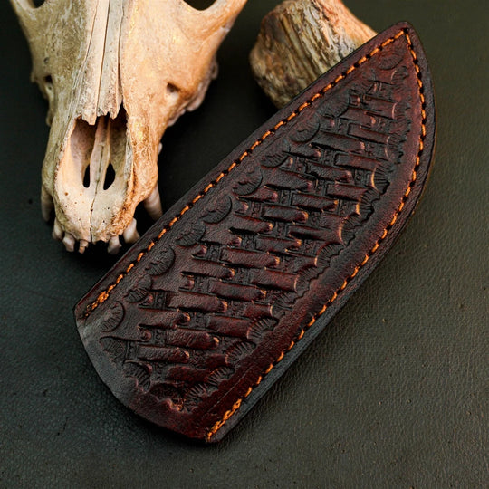 A dark brown leather knife sheath with embossed fish patterns and orange stitching, displayed on a dark textured surface next to animal bones.