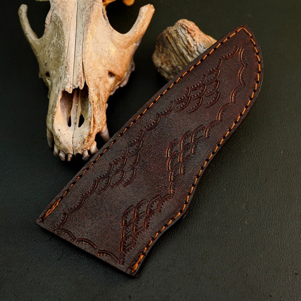 A dark brown leather knife sheath with embossed western-style stitching, displayed on a dark textured surface with a deer skull and wood prop in the background.