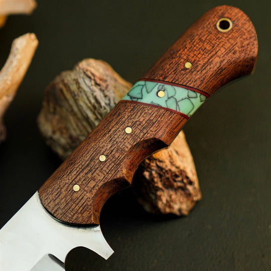 A close-up of a handcrafted knife with a rich reddish-brown wood handle, featuring a light green marbled inlay and brass pins, resting on rustic wood pieces against a dark background.