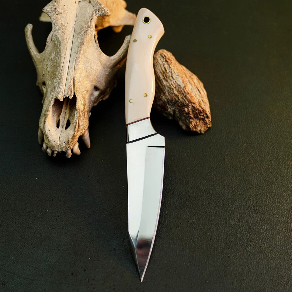 A fixed-blade knife with a polished silver steel blade and a cream-colored bone handle, displayed vertically on a dark textured surface next to a deer skull and a piece of driftwood.