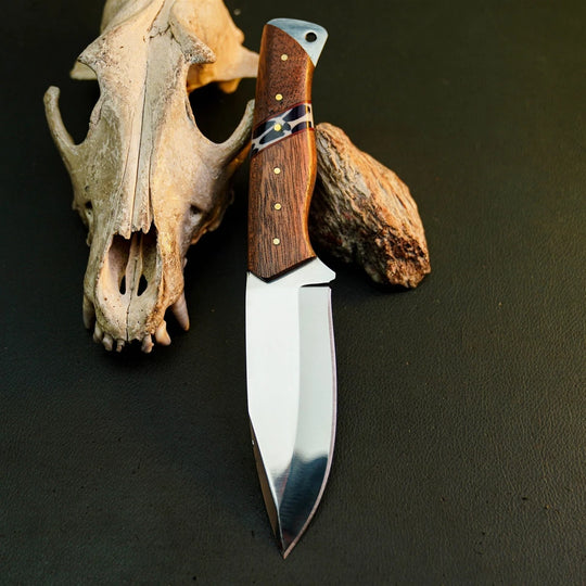 A fixed-blade knife with a polished silver blade and a brown wood-grain handle with brass pins, displayed on a dark textured surface next to a deer skull and a small rock.