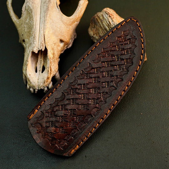 A dark brown leather knife sheath with an embossed basketweave pattern and tan stitching, displayed on a dark textured surface next to a deer skull and wood piece.
