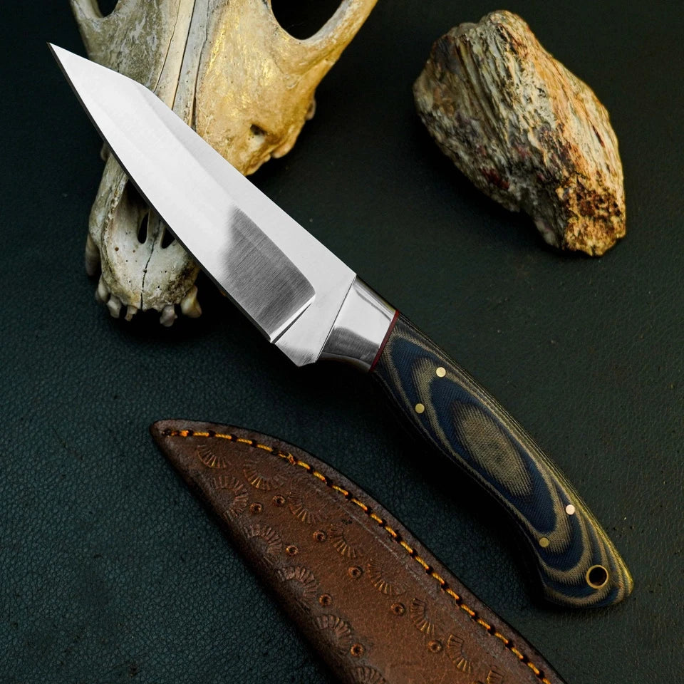 A fixed-blade knife with a satin-finished silver blade and a dark blue, gray, and brown marbled handle, displayed diagonally on a dark textured surface next to its brown leather sheath. A deer skull and a rock are used as styled props in the background.