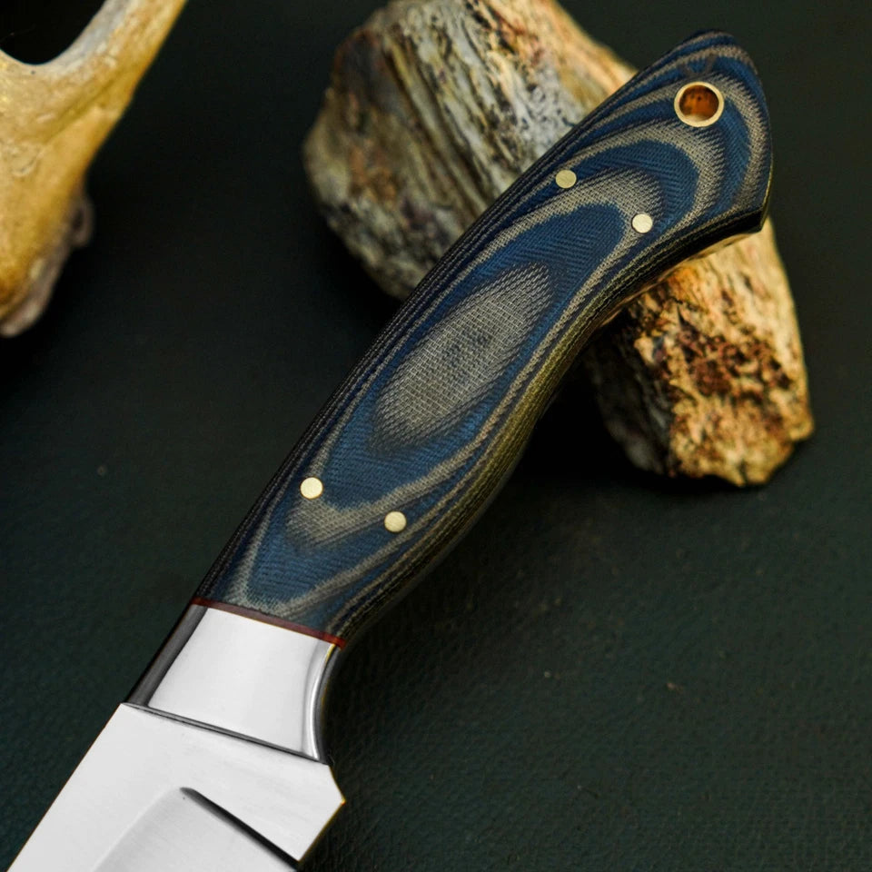 A close-up of a fixed-blade knife with a dark blue and black patterned handle and a silver steel blade, resting against natural rocks on a dark textured surface.