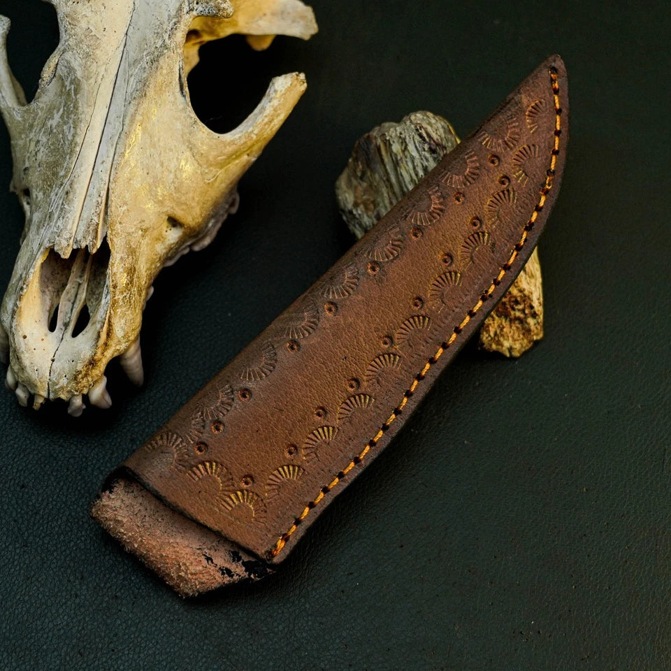 A brown leather knife sheath with an embossed fan pattern and visible orange stitching, displayed on a dark textured surface next to animal bones.